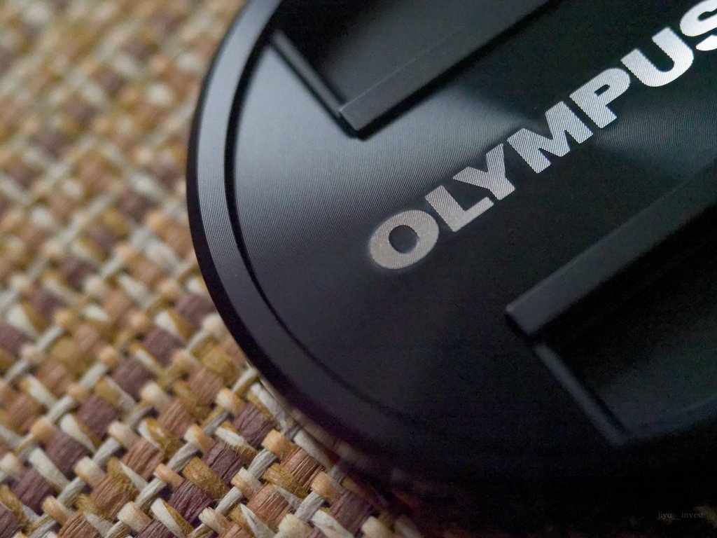 12-40mm F2.8 PROで寄って撮ったオリンパスのレンズキャップ。刻印の凹凸まで写り、背後の敷物はぼけている