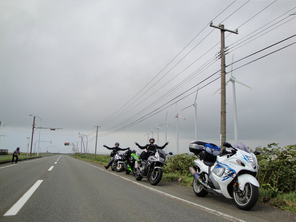 曇り空の下、風車が並ぶ道の路肩に4台のバイクを停め、ヘルメット姿の仲間が両手を広げている